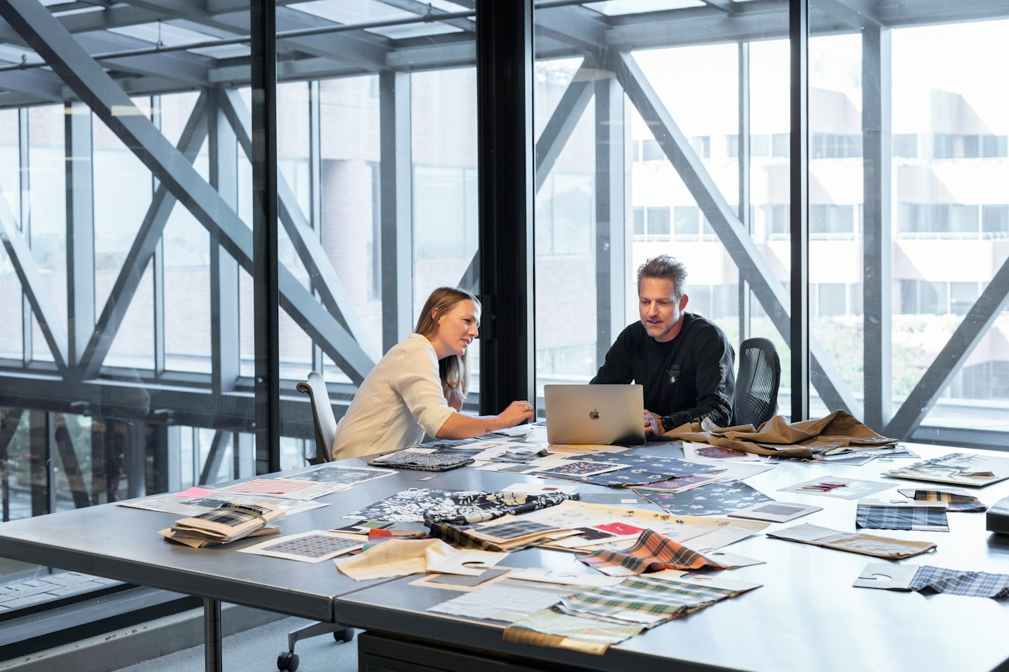 Two business people review fabric swatches at a table with a laptop.